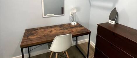A room with a wooden desk, a white chair, a dresser, a mirror, and a shelf with a lamp.