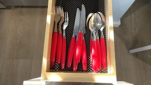 Un tiroir recouvert d’un tapis noir, contenant un couteau, une cuillère, une fourchette et une paire d’ustensiles à manche rouge.
