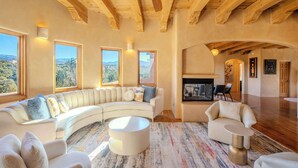 A spacious living room with a curved white sofa, a round coffee table, and a fireplace. Large windows offer a view of the outdoors. - Amazing views, Stunning Sunny Interior (Santa Fe)