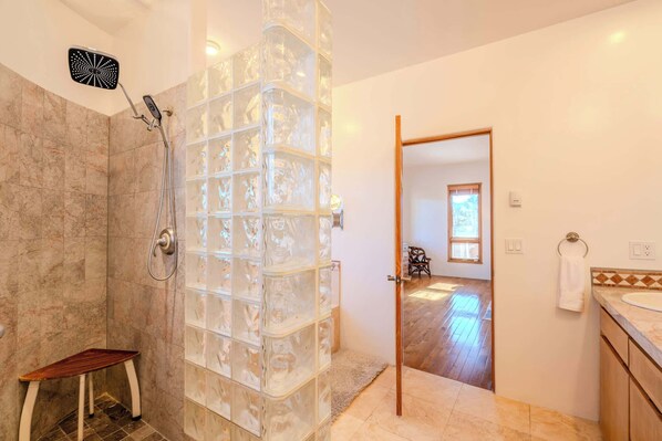 Un baño moderno con cabina de ducha de vidrio, un taburete de madera y vistas al dormitorio a través de una puerta abierta.