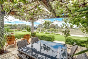 Uma área de estar coberta ao ar livre, com mesa de vidro e cadeiras de vime, cercada por vegetação e vista para um jardim.