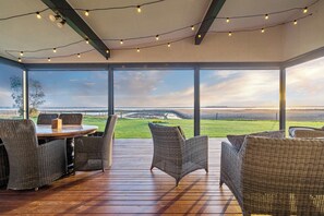Área de estar coberta ao ar livre com móveis de vime e luzes de corda, com vista para uma paisagem gramada.