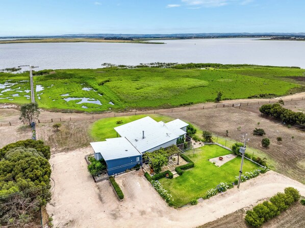 Um edifício azul com telhado plano, rodeado por campos verdes e uma grande massa de água.
