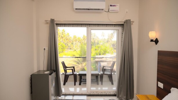 A hotel room with a bed, a desk, a chair, an air conditioner, and a view of palm trees and a thatched-roof structure outside. - Parth Guesthouse (Morjim)