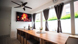 A modern kitchen with a wooden countertop, built-in seating, a ceiling fan, a flat-screen TV displaying a fireplace, and large windows with curtains.