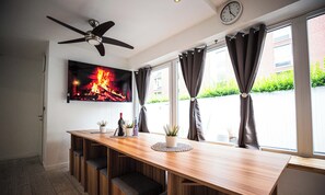 A modern kitchen with a wooden countertop, built-in seating, a ceiling fan, a flat-screen TV displaying a fireplace, and large windows with curtains.