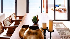A dining area with a wooden table, chairs, a bowl of fruit, candles, and a view of the ocean through large windows. - Anzhu Seamate Villa Samui (Koh Samui)
