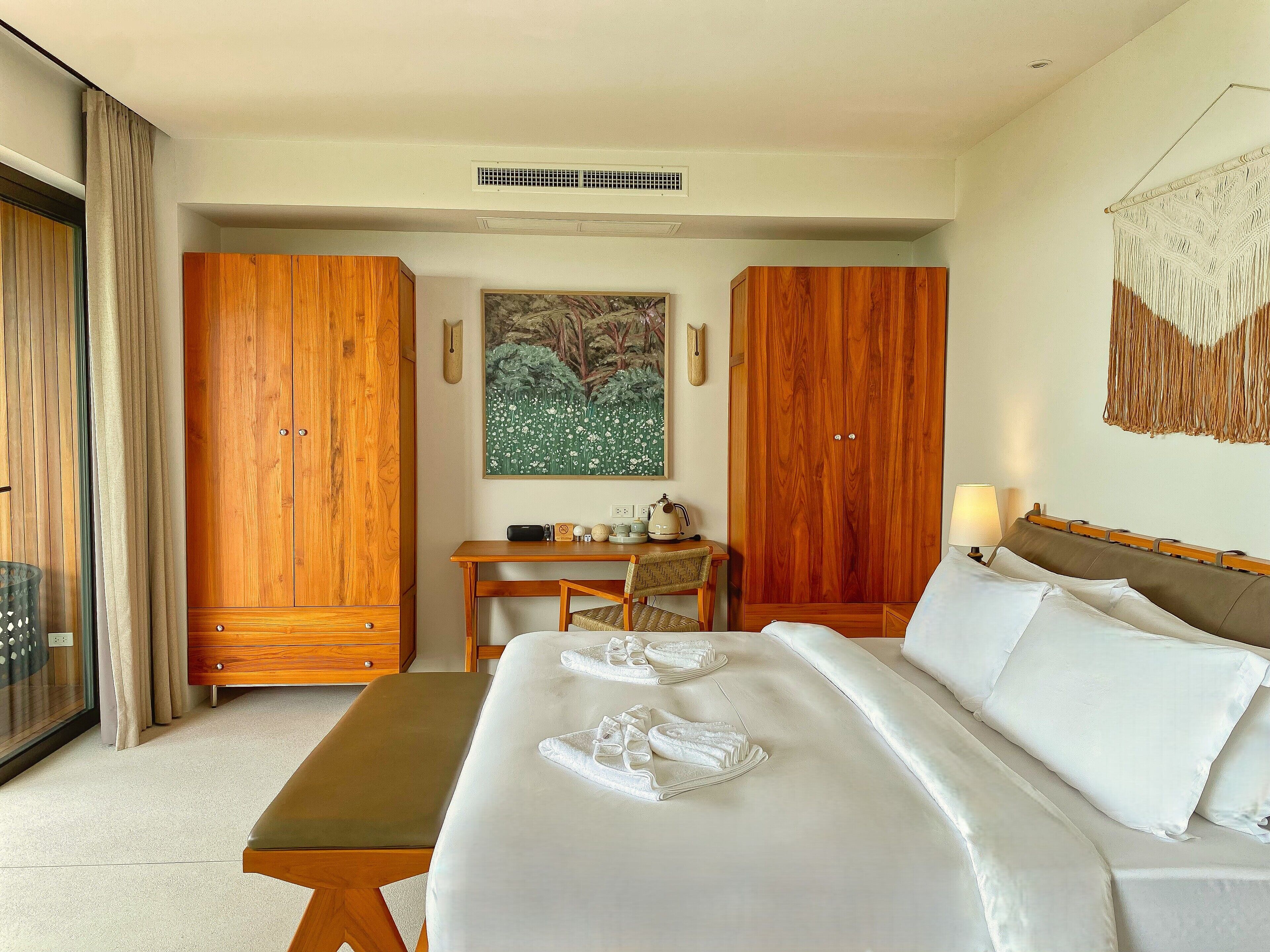 A hotel room with a large bed, wooden wardrobe, a desk with a chair, and a painting on the wall.