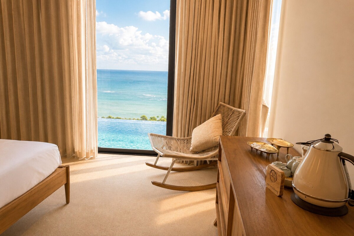 A hotel room with a bed, a rocking chair, a dining table with a kettle, and a view of the ocean.