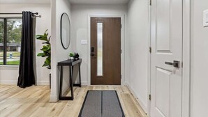 Un pasillo con piso de madera, un felpudo a rayas blanco y negro, una cortina negra, una pequeña mesa con una planta y una puerta con interruptor de luz.
