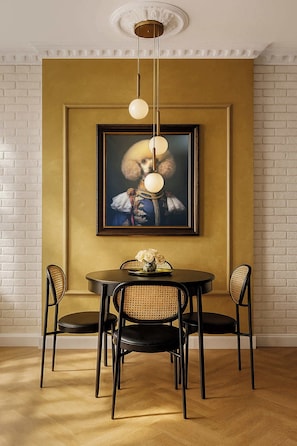 A dining area with a round table, four chairs, a framed painting, and a hanging light fixture.