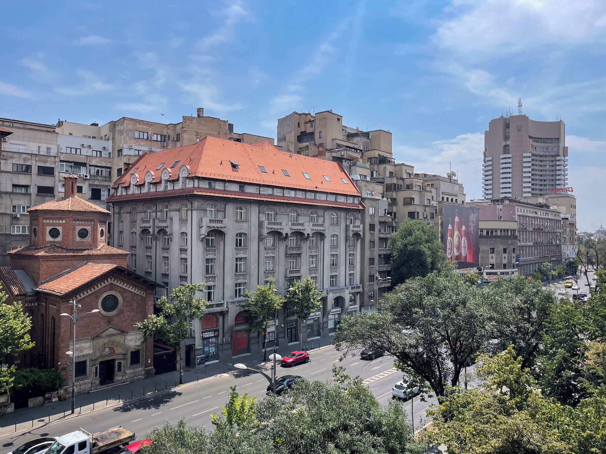 A historic building with a red-tiled roof and classical architecture, surrounded by modern buildings and a busy street.