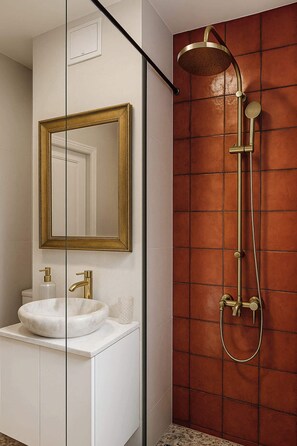 A bathroom with a white vanity, a gold-framed mirror, a gold showerhead, and a red-tiled shower wall.