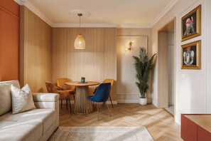 A modern living room with a round wooden table, two chairs, a sofa, a hanging light fixture, a potted plant, and framed artwork on the wall.