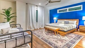 Une chambre à coucher comprenant un lit, des tables de chevet, un banc, un ventilateur de plafond et une fenêtre avec des rideaux.