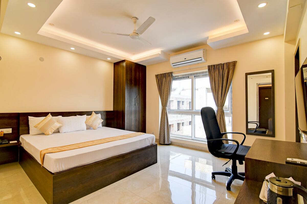 A hotel room with a large bed, a desk, a chair, a ceiling fan, an air conditioning unit, and a window with curtains.
