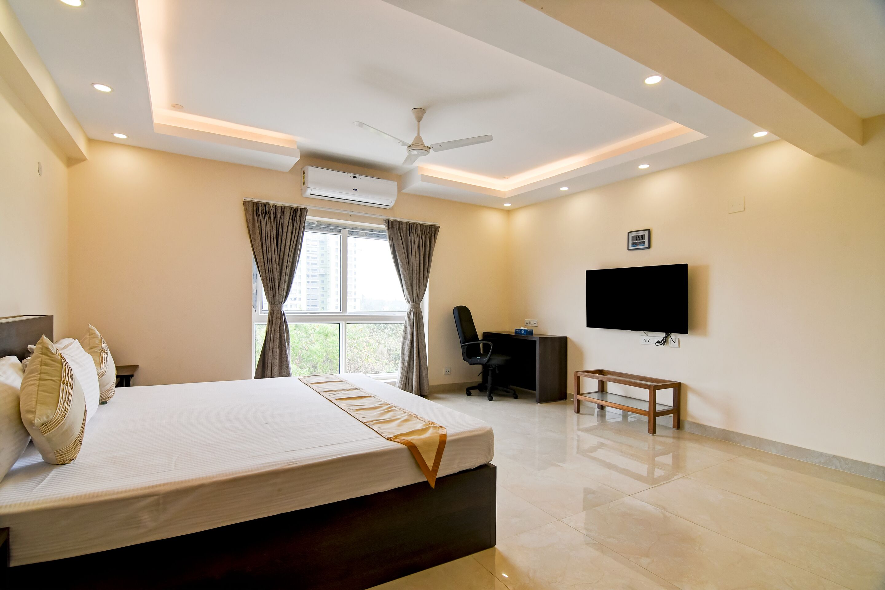 A hotel room with a large bed, a desk, a chair, a TV, and a ceiling fan.