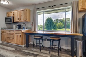 A kitchen with wooden cabinets, a built-in microwave, a window with a view of trees, and two bar stools.