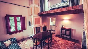 Un coin repas accueillant avec un sol en briques, une table en bois et des chaises. On y trouve une cheminée, une fenêtre avec des volets et un escalier menant à un niveau supérieur.