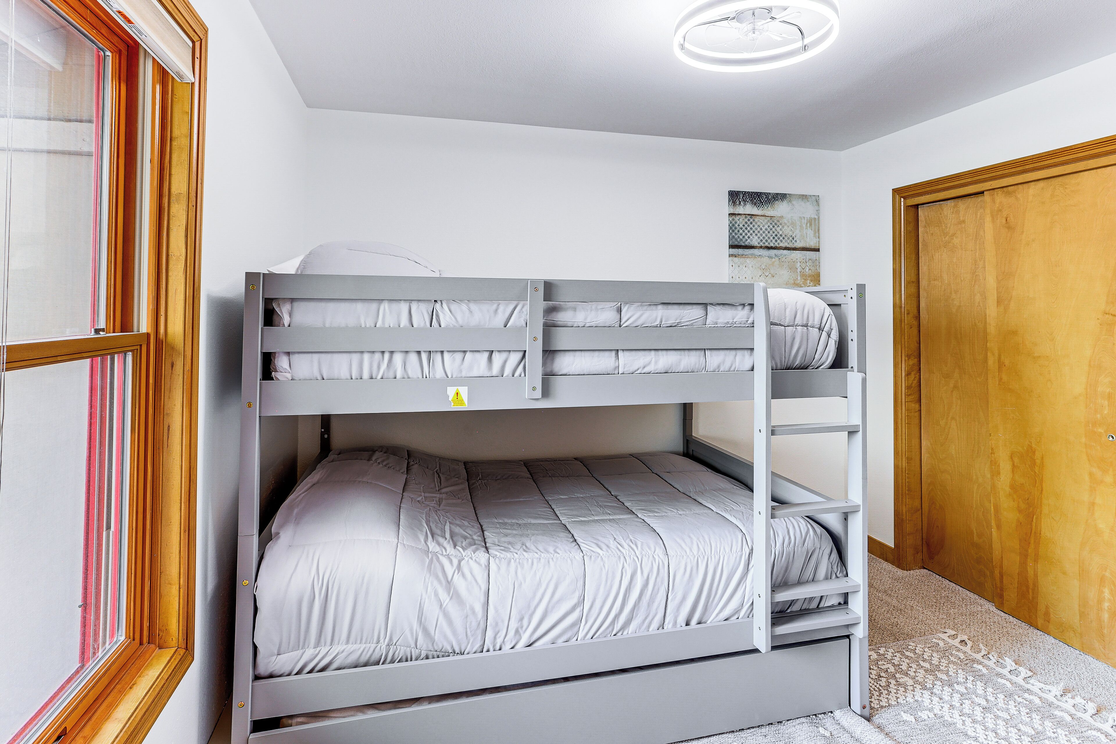 A bunk bed with a ladder, a window with blinds, a wooden door, and a ceiling light.