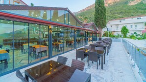 Un área de comedor al aire libre con mesas y sillas, un cerramiento de vidrio y montañas al fondo.