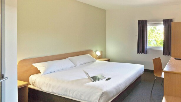 Une chambre d’hôtel avec un lit simple, un bureau, une chaise, une table de chevet et une fenêtre avec des rideaux.