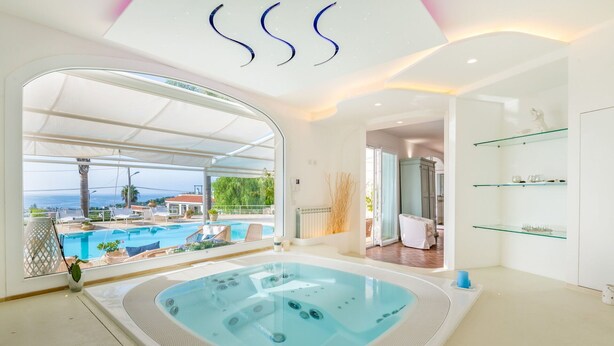 Un espace piscine intérieur moderne, doté d’un jacuzzi, offrant une vue sur la mer et comprenant une étagère en verre.