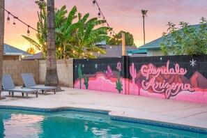 A swimming pool with lounge chairs, a mural with cacti and the text 'Gryphon Arizona,' and string lights.