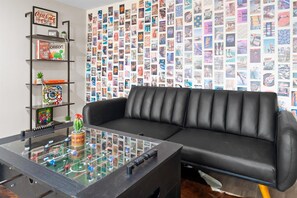 A modern living room with a black leather sofa, a glass coffee table with a board game, a bookshelf with various items, and a wall covered with a collage of images.