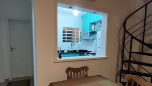 Een eethoek met een houten tafel, stoelen, een groot decoratief blad, een raam met uitzicht op een keuken en een spiltrap.