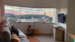 Een woonkamer met een groot raam en uitzicht op de stad, een bank, een lamp, een tafeltje met een plant en een televisie aan de muur.