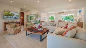 A spacious living room with a large sofa, a coffee table, and a wicker chair. There is a painting on the wall and a window providing natural light.