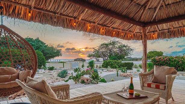 A covered outdoor seating area with wicker furniture, a table with a bottle and glasses, and a view of the sunset over the sea.