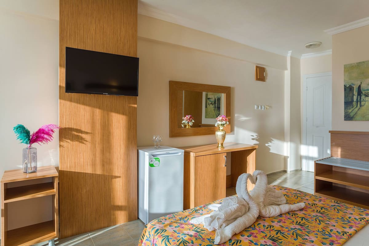 A hotel room with a bed, a wooden wardrobe, a small table with a vase and a wine glass, a refrigerator, a television mounted on the wall, and a painting on the wall.