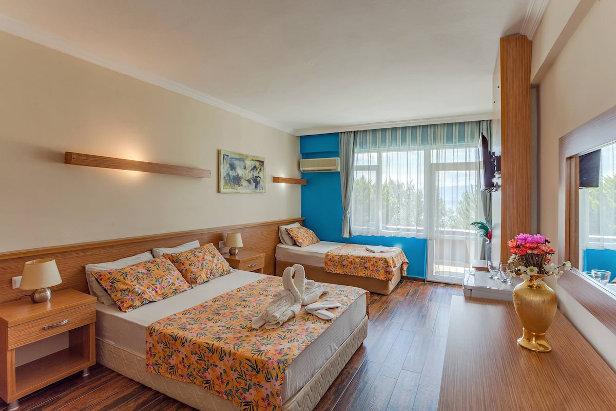 A hotel room with two beds, a wooden headboard, a bedside table with a lamp, a window with curtains, and a vase with flowers.