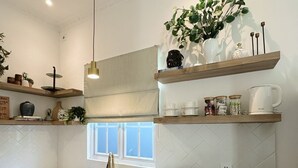 Et moderne køkken med hvide skabe, en indbygget ovn, mikrobølgeovn og vask. Der er hylder over disken med diverse køkkenartikler og en potteplante.
