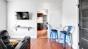 Un salon avec un téléviseur, un canapé et une table basse. On peut apercevoir une cuisine à travers une porte ouverte.