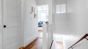 A hallway with white walls and wooden flooring leading to a living room with a couch and a window. - Quaint Escape for 2 Near Richmond Battlefield Park (Richmond)