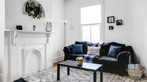 A living room with a fireplace, a black sofa with blue cushions, a coffee table, and a white rug. - Quaint Escape for 2 Near Richmond Battlefield Park (Richmond)