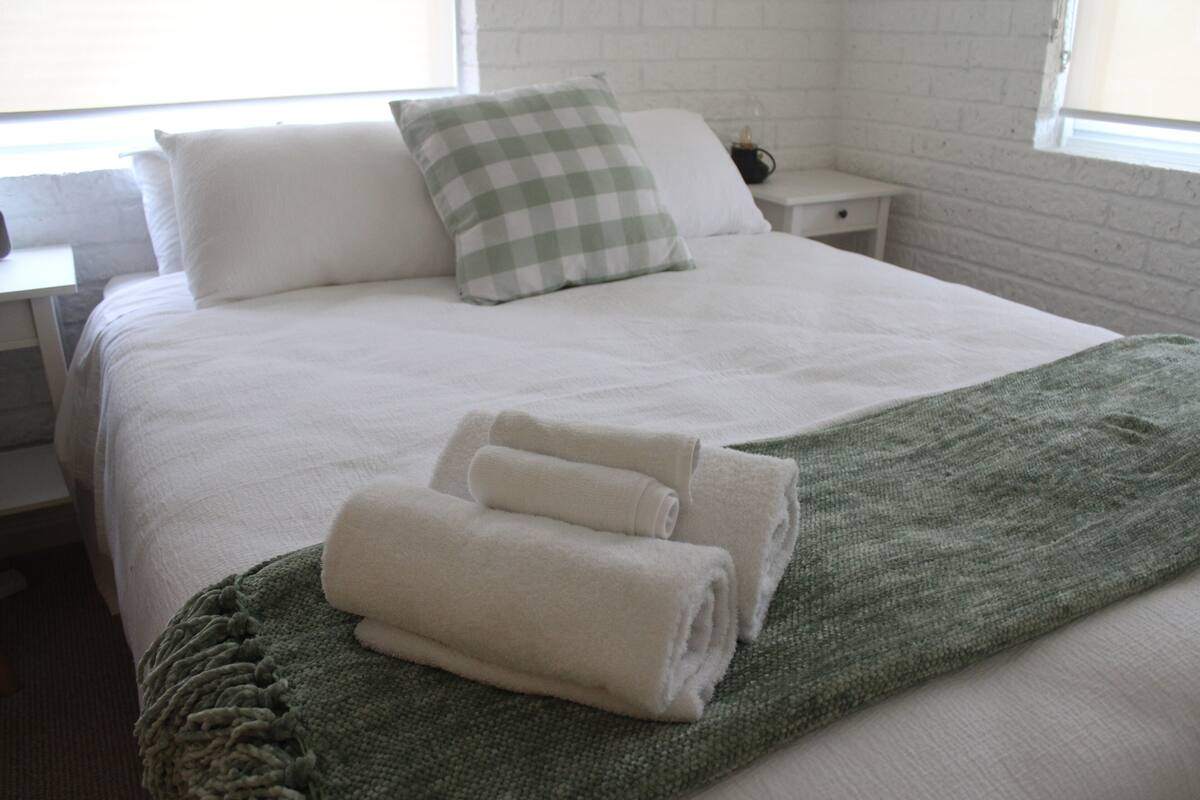 A neatly made bed with white bedding and a green throw blanket, two rolled towels, and a checkered pillow.