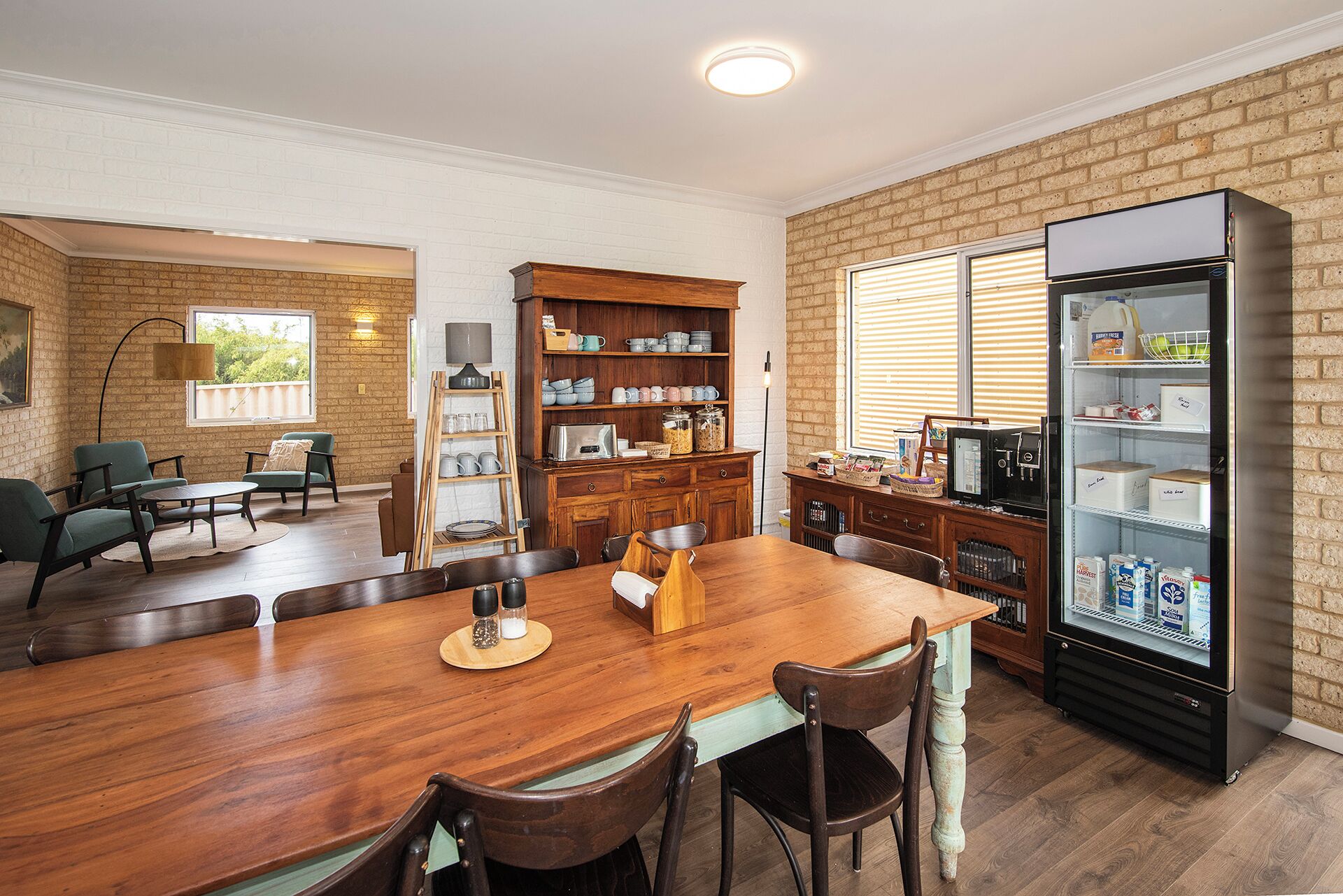 Un comedor con una mesa de madera, sillas, un refrigerador lleno de bebidas y una pared de ladrillo.