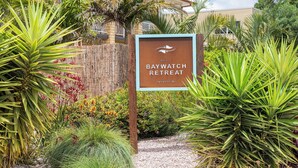 Letrero de Baywatch Retreat con valla de madera rústica y exuberante follaje.