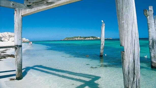 Un muelle de madera que se adentra en aguas turquesas cristalinas, con una playa arenosa y afloramientos rocosos al fondo.
