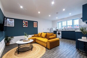 A modern living room with a sofa, coffee table, and a dining area. There is a kitchen with a washing machine and a large window.
