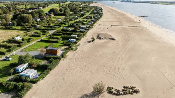 Vue aérienne d’un camping, situé près d’une plage de sable, avec des rangées de caravanes et, au loin, le profil d’une ville.