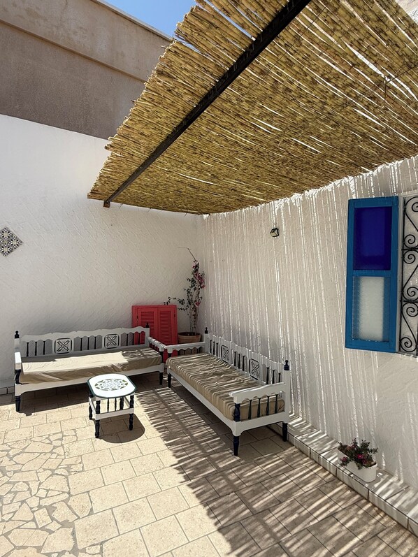 Une cour dotée d’un toit de chaume, d’un mur blanc orné d’une fenêtre bleue et d’un banc agrémenté d’un coussin à motifs.