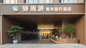 Entrada de hotel con diseño moderno, destacada por un letrero iluminado grande, plantas en macetas y un interior bien iluminado visible a través de puertas de vidrio.