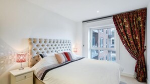 Een slaapkamer met een gestapeld hoofdbord, een bed met witte beddengoed, een nachtkastje met een lamp en een raam met uitzicht op gebouwen.