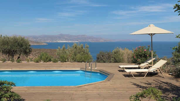 A pool with lounge chairs and an umbrella, surrounded by greenery and a view of the sea.