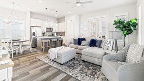 A modern living room with a grey sofa, ottoman, and a dining area with white chairs.